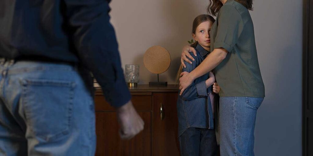 A mother protectively embraces her worried young daughter while facing a threatening man standing in the foreground indoors.